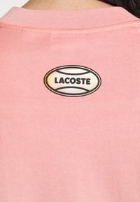 T-shirt en coton rose avec un col côtelé et un logo rond Lacoste brodé au dos en noir et blanc. Texture lisse.
