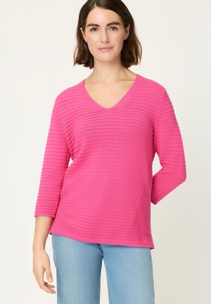 Femme portant un pull côtelé rose vif à col en V avec des manches trois-quarts et un jean bleu clair, debout devant un fond uni clair.