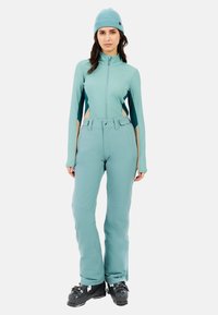 Lichtblauwe skikleding bestaande uit een getailleerde rits-top en bijpassende broek. Textuurstof met kleur-blok elementen en skischoenen.