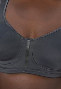 Szary sportowy biustonosz wykonany z gładkiej, elastycznej tkaniny, z dekoltem w serek, subtelnym połyskiem i metalicznym akcentem na środku przodu.