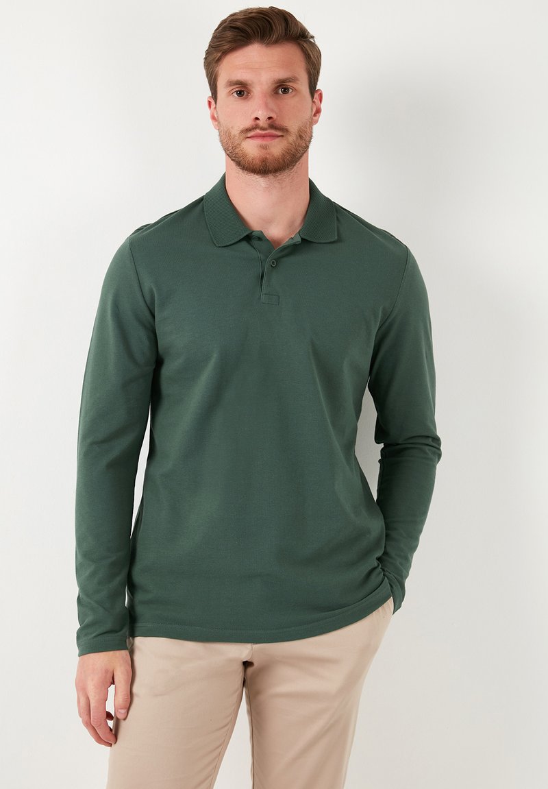 Camisa polo de manga larga en algodón verde oscuro, con cuello, dos botones y una textura suave. Combinada con pantalones beige claros.
