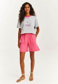 Šedé bavlněné tričko s ohrnutými rukávy a růžovým potiskem; spárováno s růžovými lněnými šortkami s páskem na stažení a skládacími lemy.