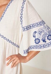 Abito di stoffa bianca con accenti ricamati in blu, caratterizzato da maniche a sbuffo e una vita arricciata. Il ricamo include motivi floreali e geometrici.