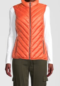 Pomarańczowa puchowa kamizelka z wysokim kołnierzem, ukośnym pikowanym wzorem oraz bocznymi kieszeniami na suwak. Nosi się ją na białą koszulę z długim rękawem i oliwkowe spodnie.