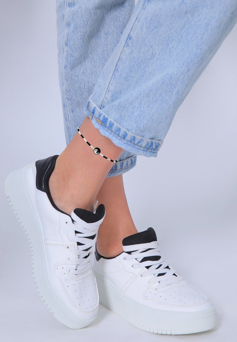 Hvide sneakers med sorte accenter, overdel i tekstureret læder og tyk hvid sål. Ankel prydet med et sort og hvidt perlesmykke med et yin-yang symbol.