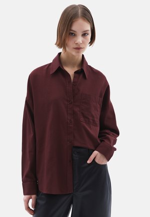 Camicia bordeaux a maniche lunghe con apertura a bottoni, colletto e una tasca sul petto. Abbinata a pantaloni neri a vita alta.