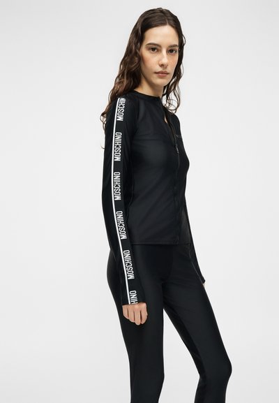 Γυναίκα με μακριά καστανά μαλλιά, ντυμένη με μαύρο τοπ και παντελόνι με φερμουάρ, με το κείμενο «MOSCHINO» επανειλημμένα τυπωμένο στα μανίκια, πάνω σε απλό φόντο.