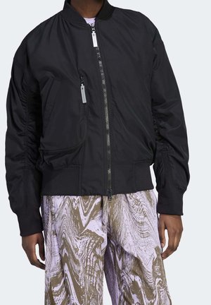 Persoon die een zwarte bomberjas met ritssluiting draagt, voorzien van een verticale ritstas, en een losse broek met bruine en lavendel gekleurde, gemarmerde patronen.