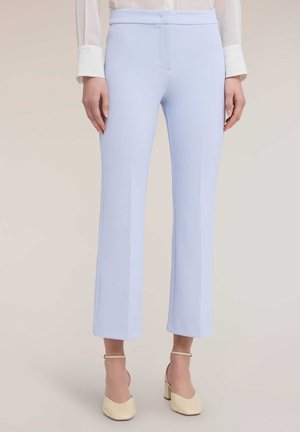 Mujer con pantalones de pierna corta a medida color azul claro con costura centrada, combinados con zapatos de tacón beige con pulsera en el tobillo y una blusa blanca transparente de manga larga.