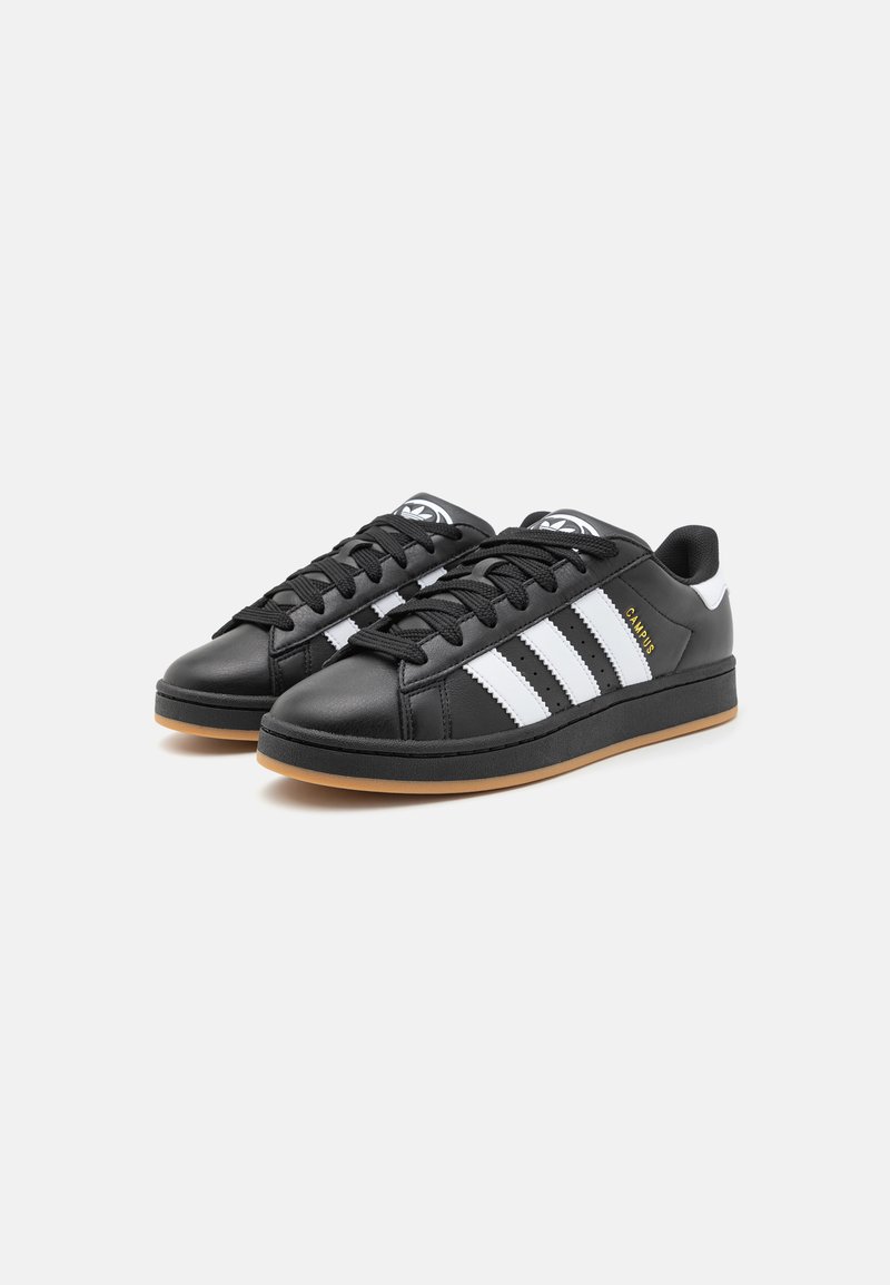 Zapatillas de deporte de cuero sintético negro con tres rayas blancas, suela de goma plana y punta redonda. Presentan cordones negros y un logo en la lengüeta.