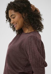Smiling woman with curly hair touching her hair, wearing a sparkly purple ribbed sweater with puffed sleeves against a plain background.