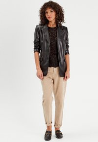 Blazer in similpelle nera sopra un top in pizzo nero, abbinato a pantaloni in velluto a coste beige e mocassini neri con dettagli in oro.
