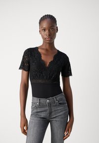 Body en dentelle noire avec des manches courtes, décolleté en V profond et bords festonnés. Associé à un jean en denim gris ajusté.
