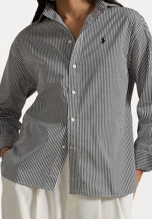 Femme portant une chemise boutonnée à rayures verticales noires et blanches avec manches retroussées et pantalon clair, mains dans les poches.