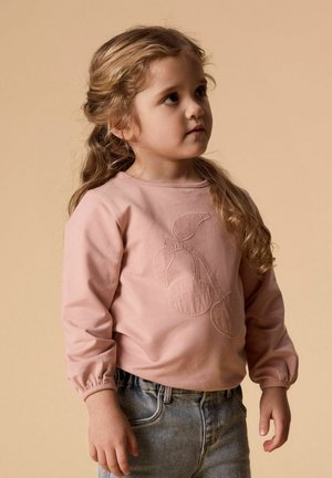 Jeune fille aux cheveux longs et ondulés portant un sweat-shirt rose clair brodé de cerises et un jean bleu, debout devant un fond beige.