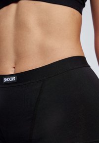 Shorts noirs taille haute en tissu doux et extensible. Ils présentent une texture lisse, des coutures plates et une taille arborant un logo.