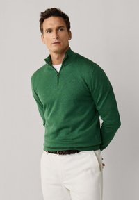 Muž, ktorý má na sebe zelený sveter so štvrťzipsom cez bielu košeľu, biele nohavice a hnedý opasok, stojí s rukami za chrbtom.