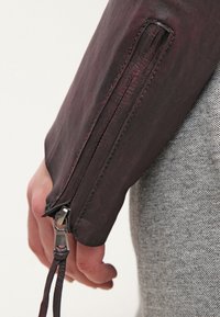 Burgundy läderjacka med dragkedja på ärmen, som har en slät yta och en justerbar rem. Grå tygbakgrund.