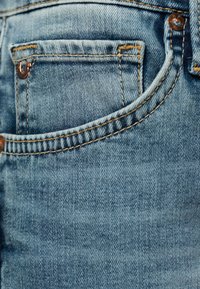 Gros plan sur la poche d'un jean en denim bleu avec des coutures orange et des rivets métalliques sur le tissu.