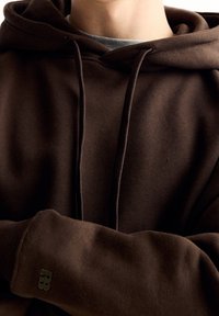 Felpa con cappuccio marrone dal finish texturizzato, con tasca a marsupio e logo discreto sulla manica. Stringhe visibili al collo.