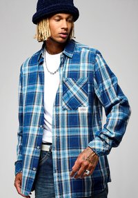 Camisa de franela a cuadros azul con bolsillo en el pecho, cierre de botones y mangas largas. Llevada sobre una camiseta blanca, con jeans de mezclilla.