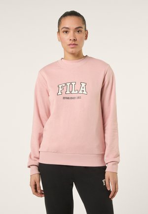 Giovane con i capelli tirati indietro che indossa una felpa rosa FILA e pantaloni neri, in piedi davanti a uno sfondo chiaro e uniforme.