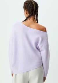 Helllila übergroßer Pullover mit schulterfreiem Design, gestrickter Textur, gerippten Bündchen und Saum. Kombiniert mit grauen Hosen.