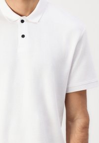 Hvid polo t-shirt med et tekstureret, ribbet design; har en standard krave og tre sorte knapper ved halsen. Korte, rullede ærmer.