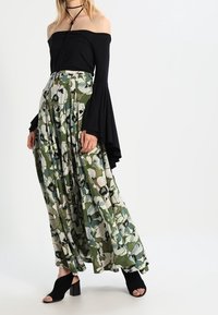 Kvinna som bär en svart off-shoulder topp med utsvängda ärmar, gröna blommiga vida byxor, svarta öppna klacksandaler och ett svart choker.