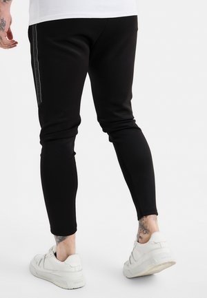 Mann trägt schmale schwarze Hosen mit seitlichem Streifen-Detail und weiße Sneakers, steht mit teilweise sichtbarem Rücken.