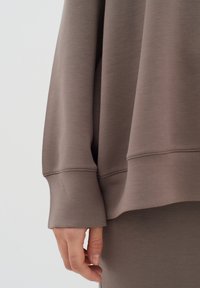 Sudadera marrón de gran tamaño con textura suave, puños acanalados y aberturas laterales, combinada con una prenda inferior ajustada a juego.