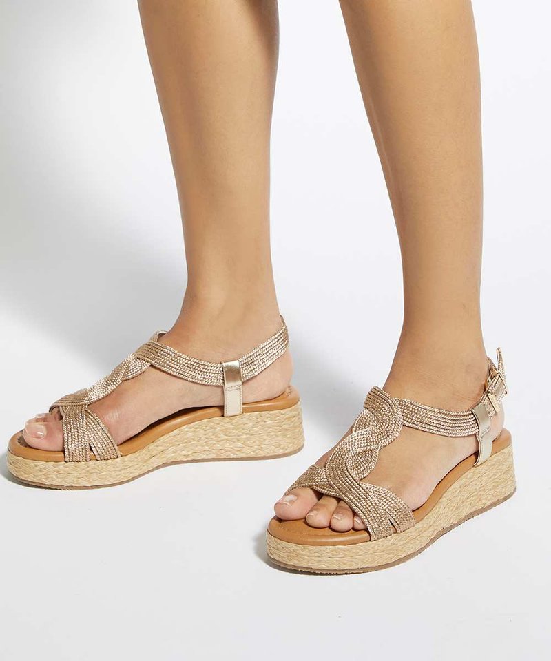 Pieds portant des sandales compensées beiges tissées avec des brides à la cheville et un motif tressé à l'avant sur fond blanc.