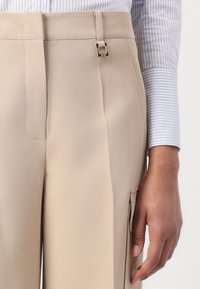 Beige kalhoty s hladkou texturou. Obsahují poutko na opasek a čtvercový kovový prvek. Viditelné detaily postranní kapsy. Část manžety pruhované košile je částečně vidět.