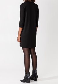 Zwart knielang jurk met driekwart mouwen, zachte stof, ronde halslijn, gedragen met zwarte enkellaarzen en doorschijnende zwarte panty’s.