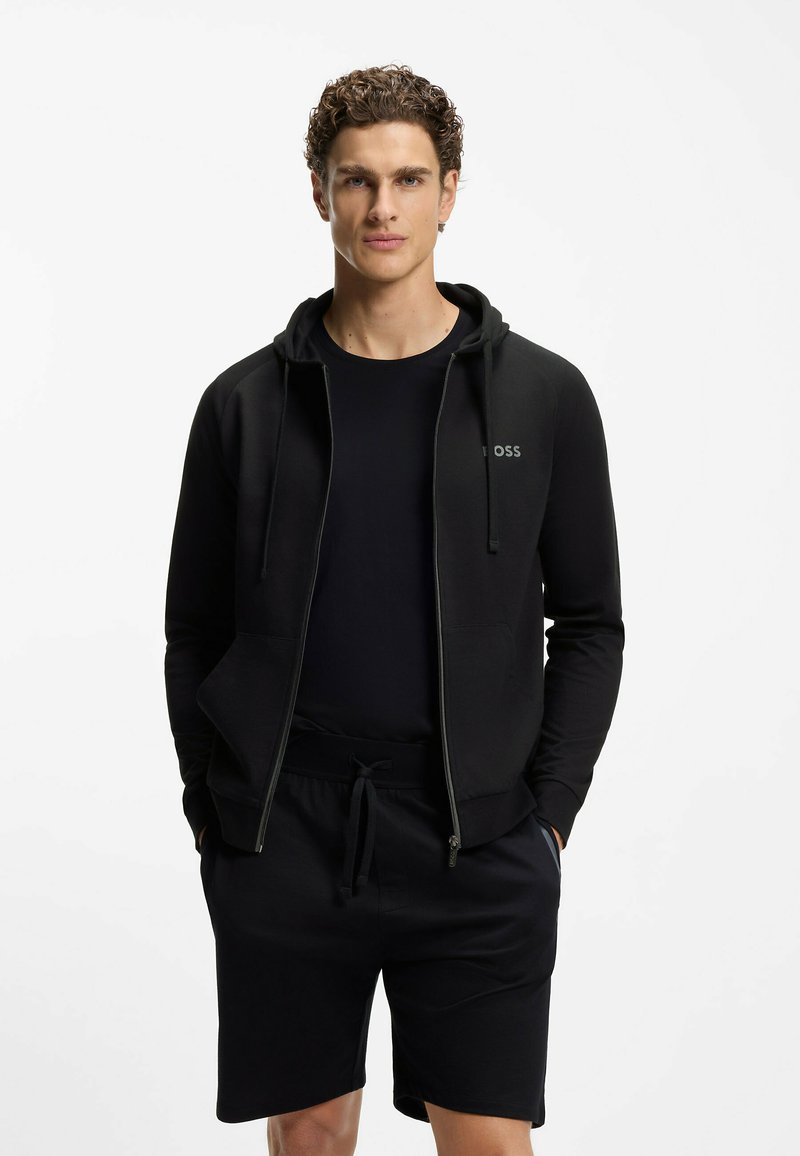 Junger Mann mit lockigem Haar trägt eine schwarze Kapuzenjacke mit Reißverschluss, ein schwarzes T-Shirt und schwarze Shorts mit Kordelzug, steht mit den Händen in den Taschen.