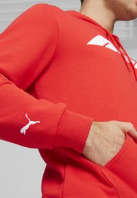 Rød bomulds hoodie med en kængurulomme, ribbede manchetter og hvidt Puma-logo på venstre ærme. Har en justerbar hætte med snore.