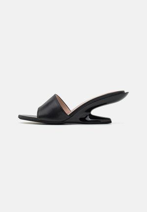 Sandalo estivo nero con una larga fascia in pelle, punta aperta e un unico tacco in plastica curvata. Texture liscia, design minimalista.