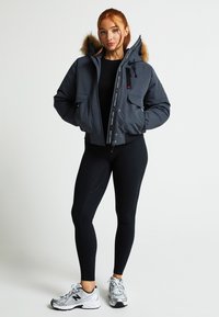 Tumši pelēkā krāsā motokrosa džaka ar kažokādas oderi kapucei. Iekļauj rāvējslēdzēju, divas priekšējās kabatas un kontrastējošus emblēmu detalizējumus. Valkāta virs melnajām legingām.