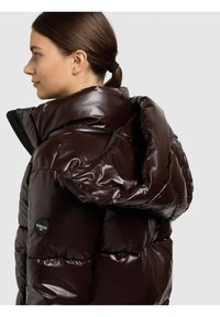 Brun vadderad jacka med glansigt ytskikt, med hög krage och dragkedjor. Inkluderar logotypdetalj på vänster sida.