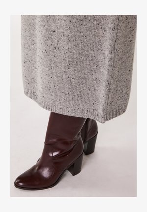 Braune Leder-Stiefelette mit quadrater Zehenform an einer Person, die einen grauen Strickrock mit strukturierten Flecken und einem gerippten Saum trägt.