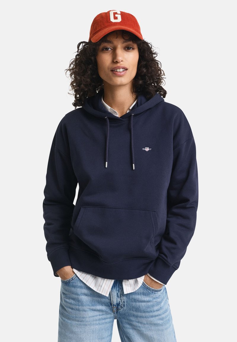 Marinblå hoodie med känguruficka, dragsko och liten logotyp. Under syns en skjorta med krage. Bärs med ljusblå jeans.