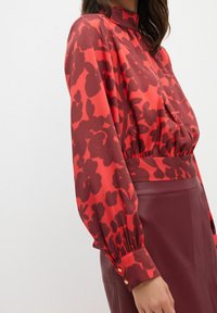 Rød bluse med iøjnefaldende blomsterprint, høj halsudskæring, samlet talje og lange ærmer med knapdetaljer. Sæt sammen med en bordeaux nederdel.