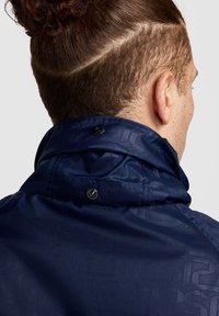 Chaqueta azul marino con un alto cuello, que presenta un patrón sutil en relieve, cierre a presión y una textura suave y brillante.
