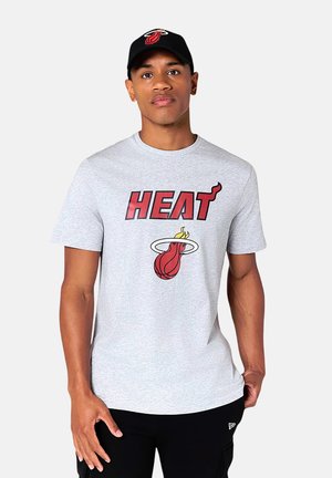 Joven con camiseta gris claro del equipo de baloncesto Heat y gorra negra de Heat, de pie con expresión neutral.