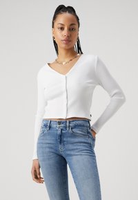 Cárdigan branco, canelado, cropped com decote em V e fecho frontal de botões. Combinado com jeans justos de um azul claro. Sem padrões ou detalhes notáveis.