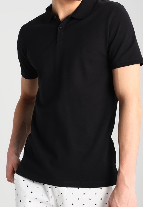Homme portant un polo noir uni à manches courtes et un short blanc avec de petits motifs en forme de losange noirs sur un fond clair.