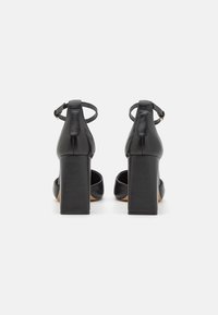 Chaussures à talons hauts en cuir noir avec talons carrés. Équipées de brides de cheville réglables et d'un design ouvert à l'avant. Texture lisse avec des accents minimes.