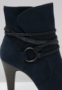 Mörkblå mocka ankestyvel med hög klack, utrustad med en flätad rem och metalldetalj runt ankeln.