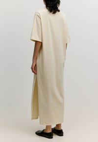 Lange beige jurk van zachte stof, met korte mouwen, een rechte snit en zijsplitten. Gecombineerd met eenvoudige zwarte schoenen.