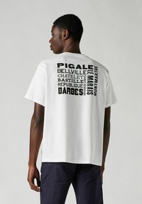 Vit bomullströja med fet svart text och stadsnamn tryckta på ryggen. Avslappnad passform med korta ärmar och rund hals.